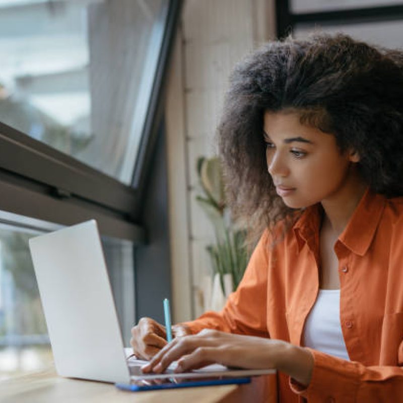 Young student using laptop computer, studying, learning language, exam preparation. Pensive African American woman freelancer writes notes working at home